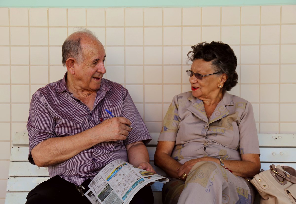 Casal de idosos sentado em um banco, conversando, ele com um jornal nas mãos