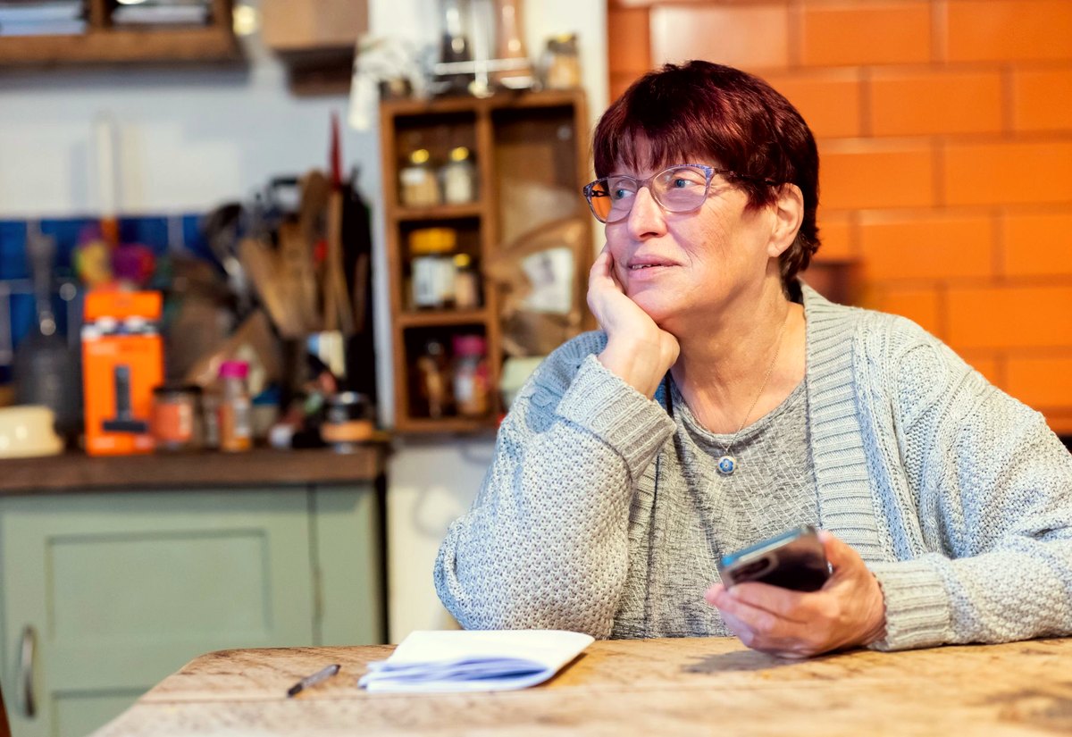 Senhora de óculos e casaco cinza, sentada à mesa da cozinha com o celular na mão, olha para longe pensativa