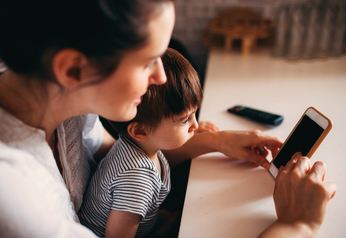 Mãe sentada à mesa mostra o celular ao filho pequeno, atenta a ele