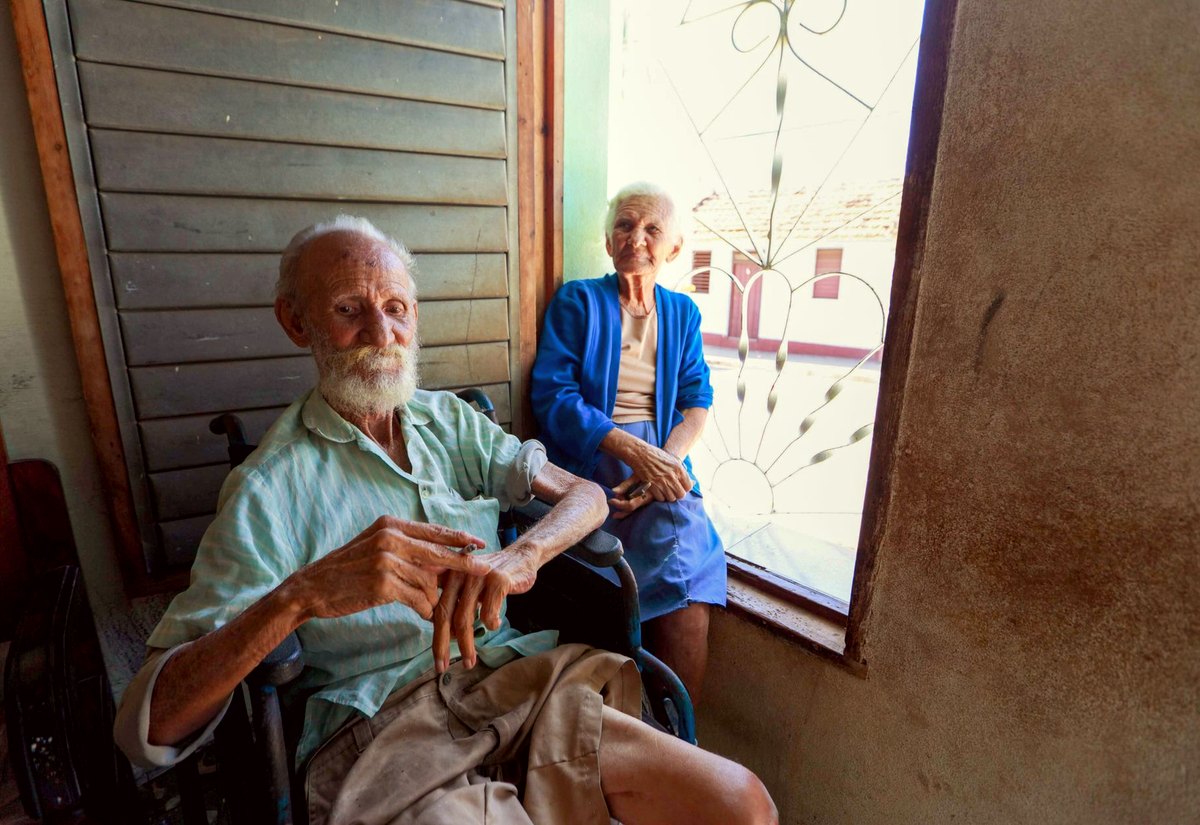 Casal idoso na sala de casa, o senhor em cadeira de rodas junto à janela