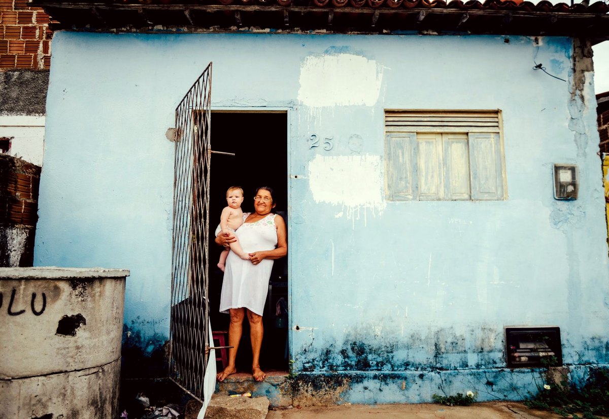Mãe com o bebê no colo na porta de uma casa simples de parede azul