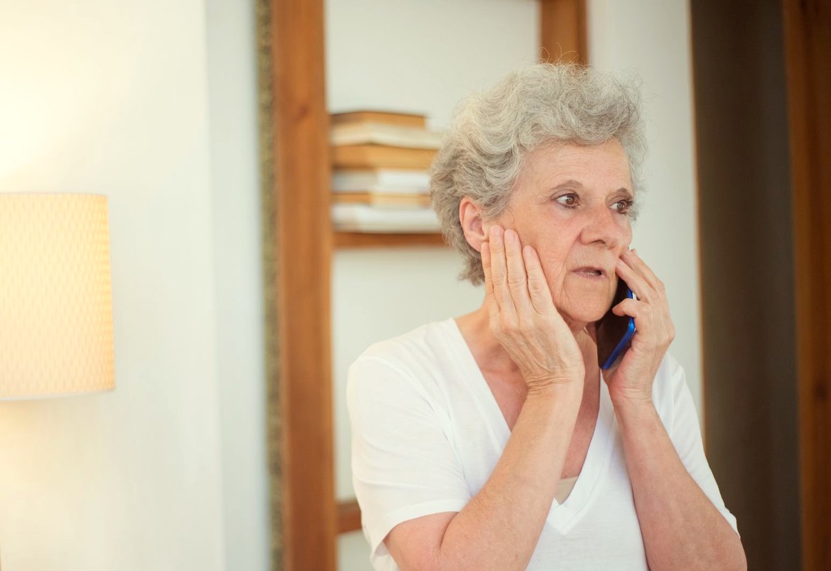 Senhora de cabelo grisalho fala ao celular com a mão no rosto, preocupada com o que acabou de ouvir