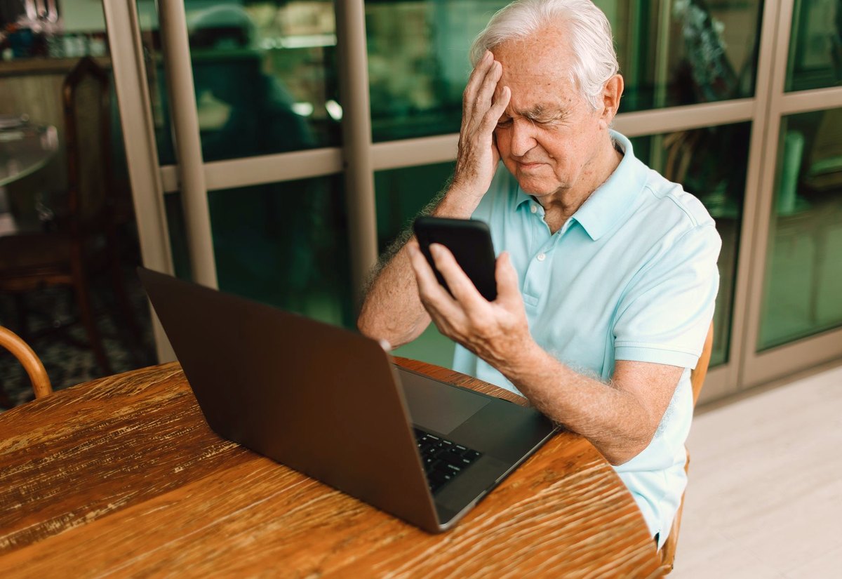 Senhor sentado no sofá com a mão na cabeça, olhando a tela do celular