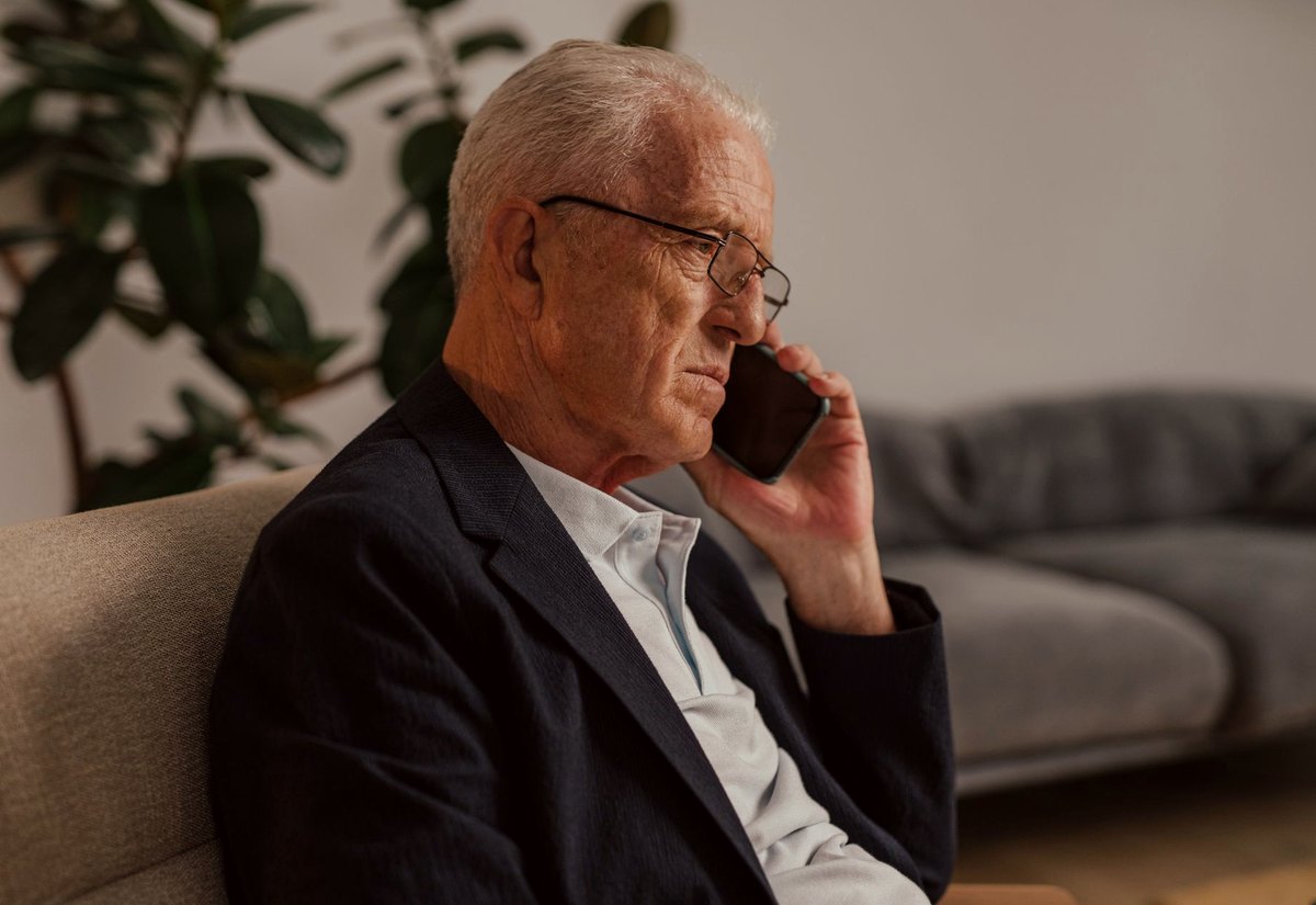 Senhor de óculos ao telefone dentro de casa, com expressão contrariada