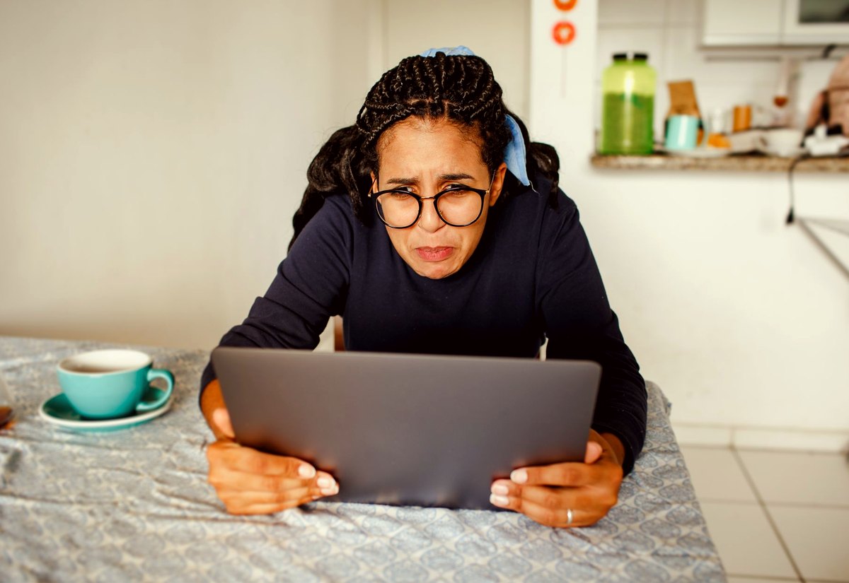 Mulher de óculos, à mesa da cozinha, olha a tela do notebook com cara de quem não reconhece o que está vendo