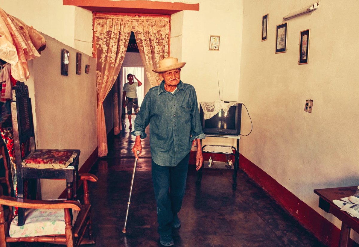 Senhor de chapéu caminha pela sala de casa apoiado numa bengala