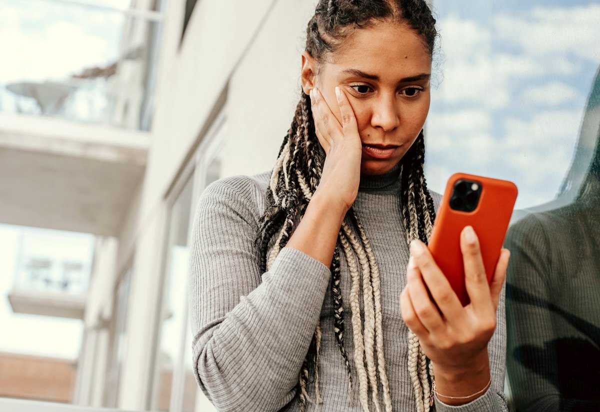 Mulher de tranças olha o celular com a mão no rosto, preocupada com o que acabou de descobrir
