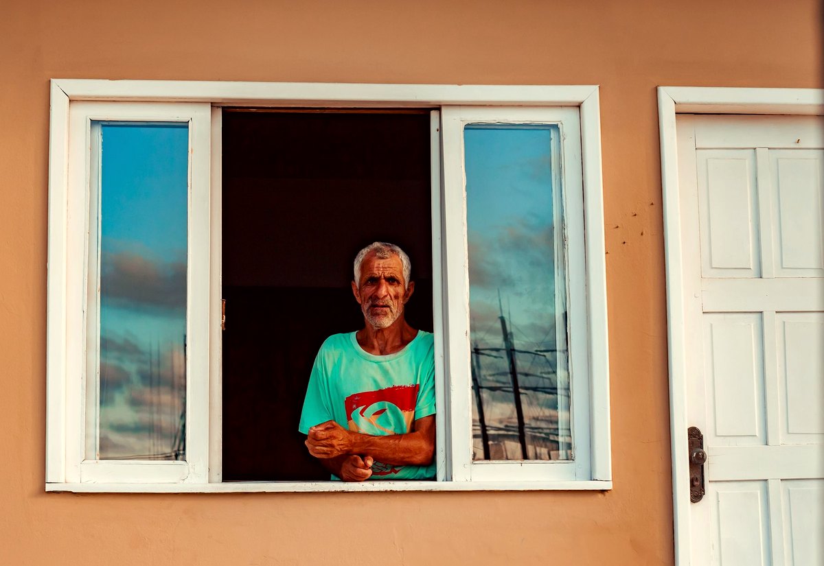 Senhor de cabelo grisalho e barba olha pela janela de casa, em Porto Belo, Santa Catarina