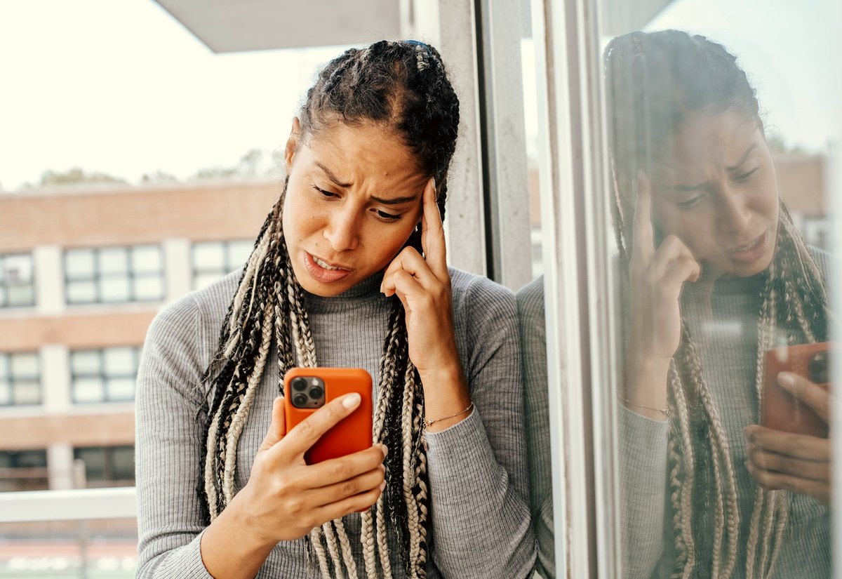Mulher encostada na janela, de sobrancelha franzida, olhando a tela do celular