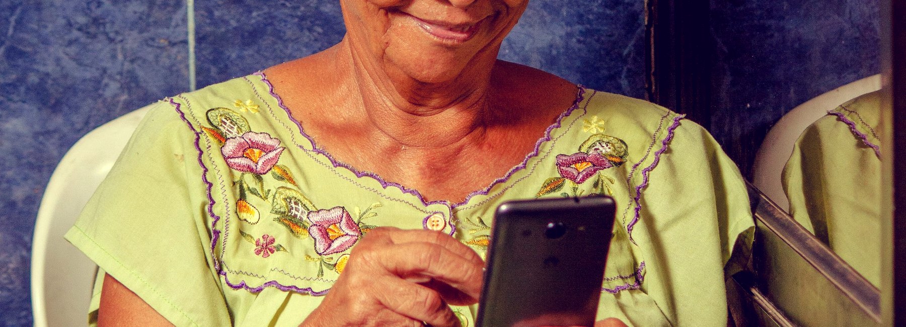 Senhora de blusa estampada mexe no celular, sentada na área de casa
